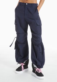 Navy cargo pants with adjustable cuffs, multiple pockets, and a relaxed fit. Worn with black sneakers featuring pink laces and patterned lining.