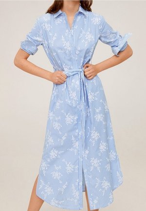 Femme portant une robe chemise à rayures bleu clair et blanches avec des motifs floraux blancs, manches retroussées et ceinture nouée à la taille.