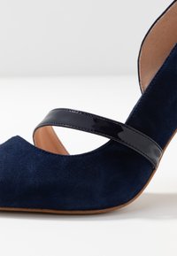 Tacco alto in suede blu navy con una fascia nera lucida sulla parte superiore e una suola color beige su uno sfondo bianco.