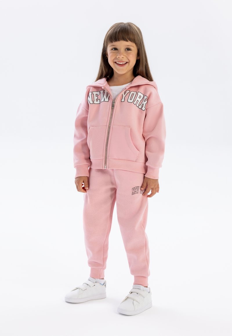 Roze zip-up hoodie met de tekst "NEW YORK", gecombineerd met bijpassende joggingbroek en witte sneakers, ontworpen voor kinderen, met een comfortabele pasvorm.
