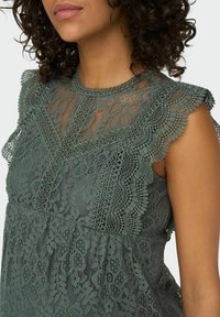 Spitzenoberteil in staubigem Grün mit gekerbten Kanten, floralen Mustern, einem hohen Halsausschnitt und geschichteten Texturen. Zartes und leichtes Design.