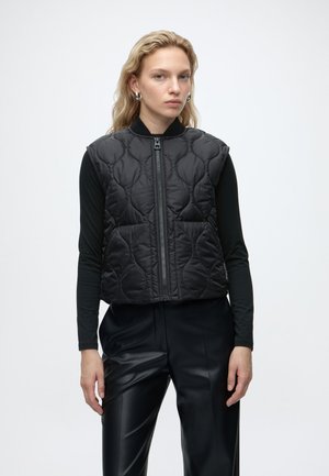 Femme blonde portant des boucles d'oreilles anneaux argentées, un gilet matelassé sans manches noir avec fermeture éclair, une chemise noire à manches longues et un pantalon en cuir noir sur fond blanc.