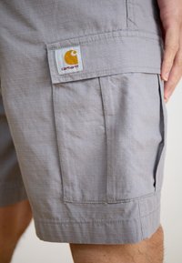 Sivé cargo šortky vyrobené z textúrovanej tkaniny majú bočnú kapsu a tkanú nášivku s logom a žltým emblémom. Ploské predné a manžetové ukončenie.