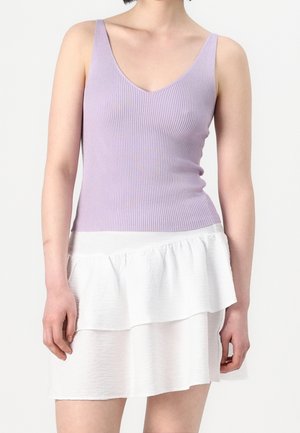 Femme portant un haut sans manches en tricot côtelé lavande avec un col en V, associé à une mini-jupe blanche à volants superposés, sur un fond uni.
