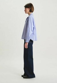 Chemise bleu clair à manches longues avec un col arrondi, associée à un jean large bleu foncé. La tenue présente des détails subtils.