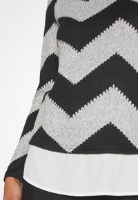 Gros plan sur un haut à manches longues à motif en zigzag noir et gris superposé sur une chemise blanche, montrant une partie de la taille et du bras.