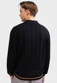 Νέος άνδρας με κοντά καστανά μαλλιά, φορώντας μαύρο υφασμάτινο πουλόβερ με καφέ ριγέ μανσέτες και γιακά, στραμμένος με την πλάτη προς το μέρος μας σε απλό φόντο.