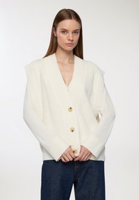 Cardigan in maglia chunky di colore bianco sporco con scollatura a V profonda, design con spalle larghe e grandi bottoni di legno, abbinato a denim.