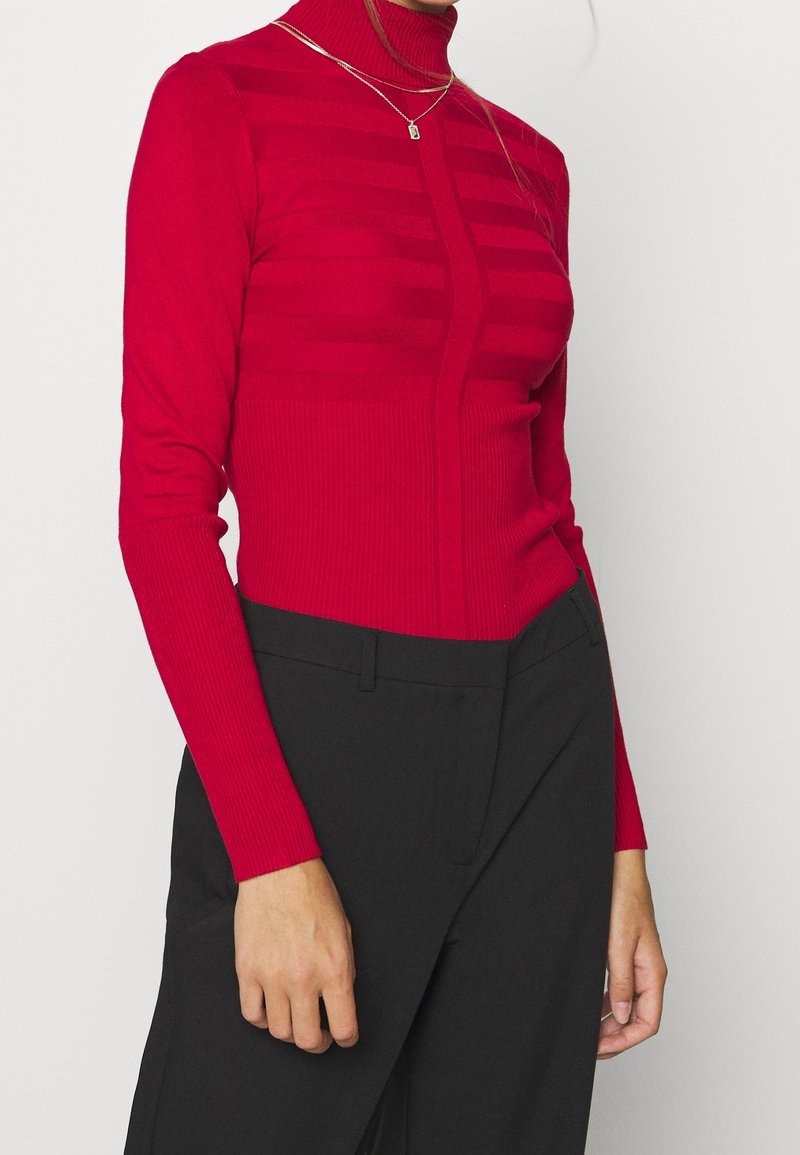 Femme portant un pull à col roulé côtelé rouge avec des rayures horizontales et un pantalon taille haute noir, debout devant un fond blanc.
