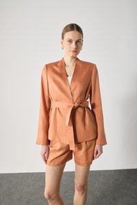 Peach-colored tailored jacket with a wrap design, long sleeves, and a tie belt, paired with matching shorts. Smooth fabric texture.