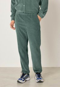 Zelene kordurojske hlače z teksturo, z elastičnim pasom in manšetami na gležnjih. V kombinaciji z sivimi športnimi čevlji.