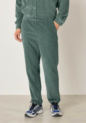 Green corduroy pants with a textured finish, featuring an elastic waistband and cuffed ankles. Paired with gray running shoes.