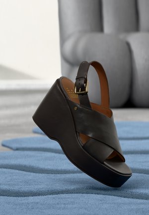 Sandale compensée en cuir noir avec brides croisées et bride à boucle à la cheville sur un tapis bleu texturé.