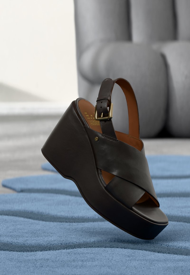 Μαύρο δερμάτινο πλατφόρμα πέδιλο με σταυρωτούς ιμάντες και πόρπη στον αστράγαλο, πάνω σε μπλε υφασμένο χαλί.