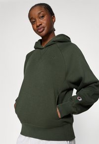 Hanorac cu glugă de culoare verde măsliniu, cu un croi lejer, având buzunar tip kangaroo, manșete din țesătură ribbed și un logo mic pe piept și pe mânecă.