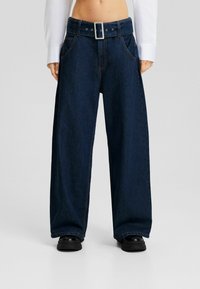 Vidvuxna mörka denimjeans med hög midja och ett stort bälte med silverfärgad spänne. Har framfickor och kontrastsömmar.