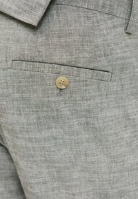 Tela de lino gris que muestra un primer plano de un bolsillo con un detalle de botón beige. La textura parece ligeramente tejida con un acabado natural.