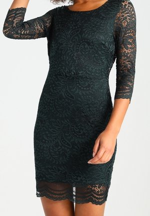 Femme portant une robe noire en dentelle ajustée avec des manches trois-quarts et des motifs floraux, debout devant un fond clair uni.