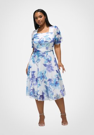 Donna che indossa un vestito midi bianco con motivi floreali blu, maniche corte a volant e sandali argentati con tacco alto e cinturini, in piedi davanti a uno sfondo bianco.