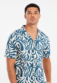 Camisa de botones de manga corta en color crema con un patrón floral abstracto en azul. La tela parece ligera y tiene un corte relajado. Botones de madera adornan la parte delantera.
