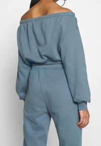 Modrá mikina bez ramínek s nařaseným výstřihem a dlouhými rukávy, v kombinaci s tepláky na gumu v pase, obě vyrobené z měkké látky.