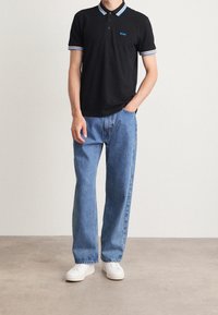 Homme portant un polo noir avec des bordures bleues et blanches, un jean bleu et des baskets blanches, adossé à un mur uni.