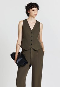 Olivgrünes ärmelloses Top mit V-Ausschnitt, fünf schwarzen Knöpfen und taillierter Passform. Kombiniert mit passender Hose; hält eine schwarze Clutch.