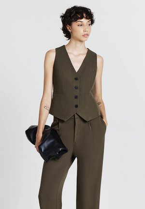 Olivgrünes ärmelloses Top mit V-Ausschnitt, fünf schwarzen Knöpfen und taillierter Passform. Kombiniert mit passender Hose; hält eine schwarze Clutch.