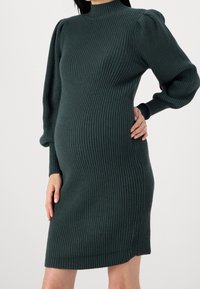 Robe en tricot côtelé de couleur bleu canard, avec un col haut et des manches bouffantes. Coupe près du corps avec un tissu texturé et longueur mi-cuisse.