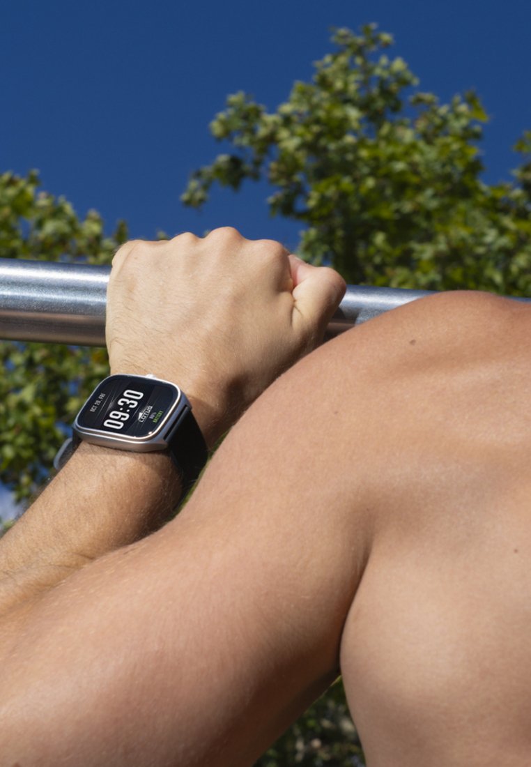 Muscular arm wearing a fitness smartwatch gripping an outdoor pull-up bar with green trees and clear blue sky in the background.