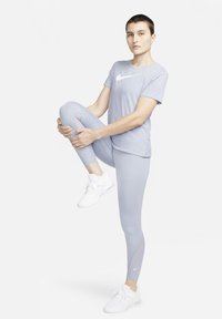 T-shirt sportivo di un leggero viola e leggings coordinati con una superficie liscia. Logo Nike bianco sulla maglietta. Modella in posa con una gamba sollevata.