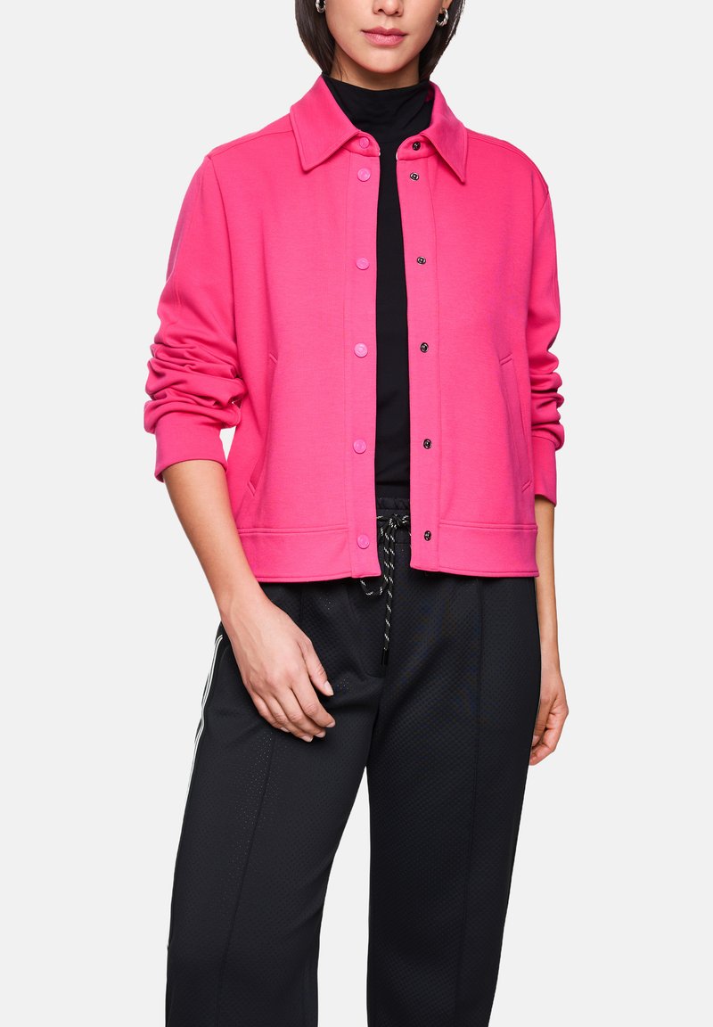 Eng anliegende, rosa Sweatshirtjacke mit durchgehendem Knopfverschluss, spitzem Kragen, Seitentaschen und hochgekrempelten Ärmeln, kombiniert mit schwarzen, zulaufenden Hosen.