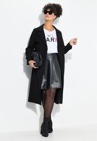 Schwarzer Trenchcoat über einem weißen T-Shirt mit Grafik, kombiniert mit einem schwarzen Lederröcken. Die Accessoires umfassen eine Sonnenbrille und eine schwarze Tasche. Schwarze Absatzstiefel.