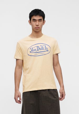 Joven de pie con camiseta amarilla clara con logo azul "Von Dutch California" y pantalones anchos oscuros sobre un fondo liso.