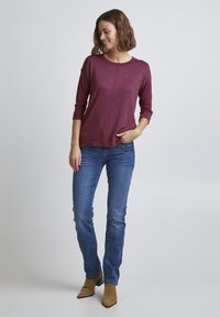 Burgundy långärmad topp med rundad halsringning, sidoknappar, i kombination med blå jeans och bruna ankelstövlar, på en neutral bakgrund.