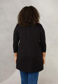 Tunika nera con maniche lunghe, a taglio dritto, di lunghezza media, con texture liscia e orlo arrotondato, indossata sopra jeans in denim blu.