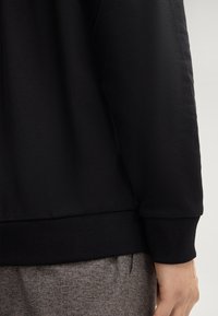 Maglione nero in tessuto testurizzato, con polsini e orlo ribattuti, e una finitura liscia sul corpo. Pantaloni grigi con motivo sono parzialmente visibili.