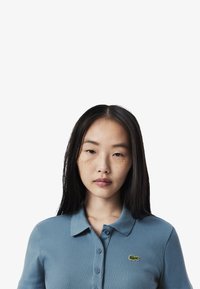Blå ribbade polo t-shirt med krage, sex knappar och en grön broderad logotyp på vänster sida. Slät textur.