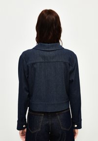 Chaqueta de mezclilla en azul oscuro, diseño corto con cuello, puños abotonados y detalles de costura visibles. La parte trasera revela una textura suave.