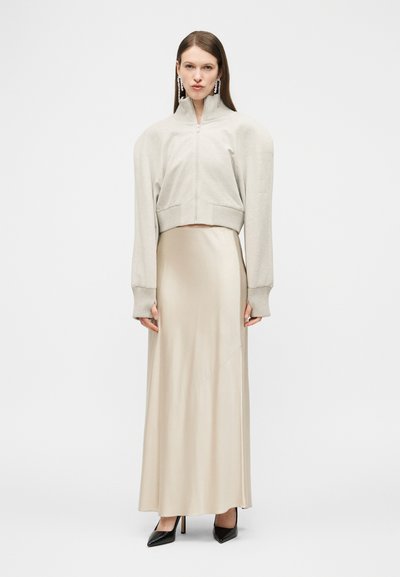 Femme portant une veste beige zippée aux épaules exagérées, une longue jupe en satin crème, des talons noirs et de longues boucles d'oreilles pendantes, se tenant droite.