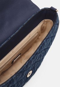 Modrá prošívaná kabelka s texturovaným vnějším materiálem, vyznačující se béžovou podšívkou, vnitřní kapsou na zip a zlatým magnetickým zapínáním.