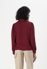 Bluză burgundy cu mâneci lungi tip puff și guler, cu o textură subtilă. Purtați cu pantaloni bej, largi.