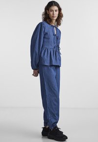 Μπλε denim σύνολο με άσπρες κάθετες ρίγες, περιλαμβάνει μπλούζα peplum, φαρδιά παντελόνια και λεπτομέρειες με κορδόνια. Συνοδεύεται από μαύρα αθλητικά παπούτσια.