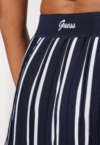 Jupe plissée marine avec des rayures verticales blanches, dotée d'une taille côtelée marquée du logo "Guess" en broderie blanche.