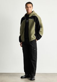 Fleece bunda na zip v olivově zelené a černé barvě, s bílými lemy a kapucí, v kombinaci s černými cargo kalhotami a černými botami.