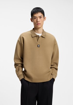 Mann trägt ein beigefarbenes Langarm-Poloshirt mit dem "HUGO"-Logo auf der Brust und schwarze Hose, steht mit den Händen in den Taschen vor weißem Hintergrund.