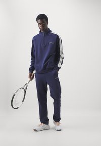 Survêtement de sport bleu marine avec col zippé, bandes latérales blanches et logo. Pantalons fuselés, assortis à des baskets blanches et une raquette de tennis.