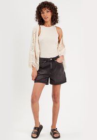 Zwarte denim shorts met een hoge taille, gecombineerd met een beige tanktop en een gestructureerde, opengehaakte cardigan met franjes. Zwarte sandalen.