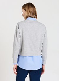 Sweatshirt grigio corto in morbido tessuto, con maniche lunghe e design semplice. Indossato sopra una camicia colletto azzurro, con pantaloni blu navy.