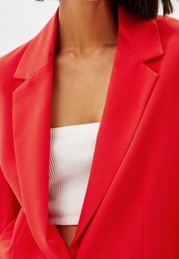 Punainen bleiseri kauluskäänteillä, sileä pinta, valkoisen ribbineuletopin päällä. Bleiserissä on muotoillut hartiat ja istuva malli.